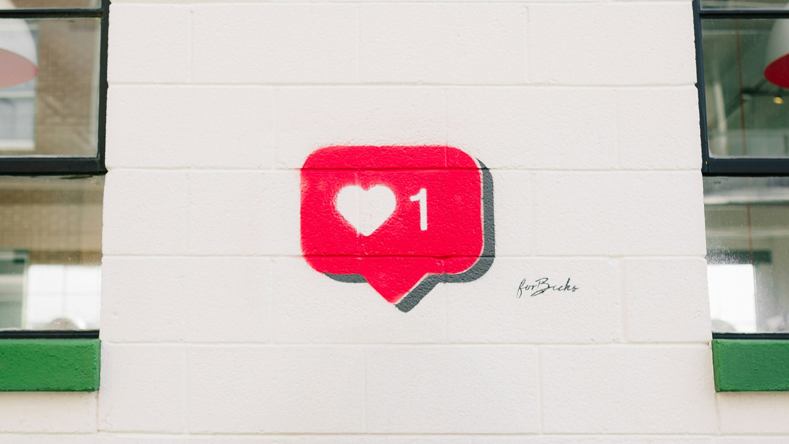Uma parede branca exibe um grafite de um ícone de notificação de 'curtida' do Instagram: um balão de diálogo vermelho com um coração branco no centro e o número '1' ao lado. A foto é usada para ilustrar um post sobre design pra redes sociais. Ao lado do grafite, nota-se a assinatura do artista em caligrafia cursiva e um tanto difícil de decifrar, possivelmente o nome é 'forBricks'.
